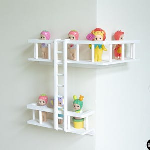 Op de afbeelding: Een witte hoekplank met twee platforms verbonden door een ladder. De planken tonen kleine, kleurrijke beeldjes, waaronder aardbei-, leeuw- en cactusontwerpen. De unit is ontworpen om perfect in een hoek te passen.