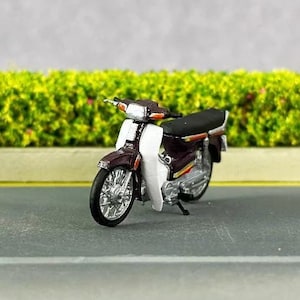 May include: A burgundy and white miniature motorcycle model with a black seat. The motorcycle is on a gray asphalt road with a green bush in the background.