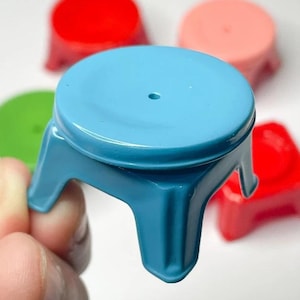 May include: A small, light blue plastic stool is held in a hand, with other colorful stools in the background. The stools have a round seat and three legs. The other stools are red, pink, and green.