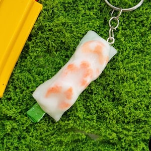 Puede incluir: Un llavero de rollo de primavera blanco y naranja con un tallo verde, unido a un llavero plateado. El llavero está sobre una superficie de musgo verde. El texto "Infinite Builds" es visible en la esquina inferior derecha de la imagen.