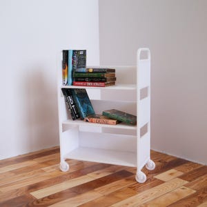 May include: White rolling bookshelf with four shelves, filled with books. The top shelf has a stack of books with the title "STEPHEN KING" visible. The second shelf has a stack of books with the title "DUNE" visible. The third shelf has a stack of books with the title "IT" visible. The bottom shelf is empty.