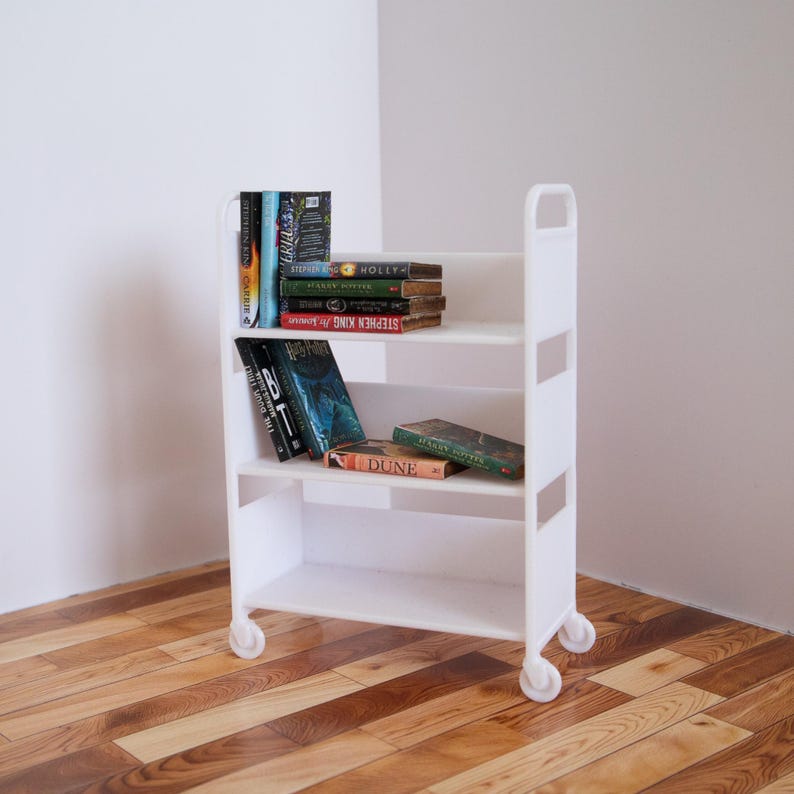 May include: White rolling bookshelf with four shelves, filled with books. The top shelf has a stack of books with the title "STEPHEN KING" visible. The second shelf has a stack of books with the title "DUNE" visible. The third shelf has a stack of books with the title "IT" visible. The bottom shelf is empty.