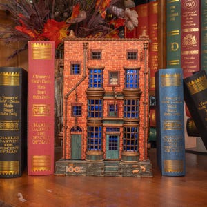 Pode incluir: Um modelo decorativo de um edif&iacute;cio com janelas azuis e fachada de tijolo, ladeado por livros de capa dura com letras douradas. Os livros incluem t&iacute;tulos como "A Treasury of World's Classics" e "Charles Darwin The Descent of Man."