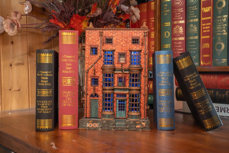 Pode incluir: Um modelo decorativo de um edif&iacute;cio com janelas azuis e fachada de tijolo, ladeado por livros de capa dura com letras douradas. Os livros incluem t&iacute;tulos como "A Treasury of World's Classics" e "Charles Darwin The Descent of Man."