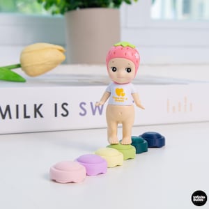 Puede incluir: Una pequeña figura con un sombrero en forma de fresa y una camiseta blanca con el texto "Time for a Break". La figura está de pie sobre una fila de objetos redondos y coloridos. Un tulipán amarillo y un libro están en el fondo.