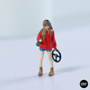 May include: A miniature figurine of a person with long brown hair wearing teal headphones, a bright red long-sleeve shirt, and blue shorts. The figure carries a green shoulder bag and holds a black steering wheel. The text Infinite Builds is visible.
