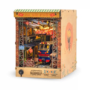 K&ouml;nnte beinhalten: Ein h&ouml;lzernes B&uuml;cherregal mit einem detaillierten Diorama einer vietnamesischen Stra&szlig;enszene mit einem Zug. Die Szene umfasst Ziegelmauern, Laternen und den Text "HA NOI PHO". Das B&uuml;cherregal hat einen Lichtschalter und eine dekorative Seitenwand.