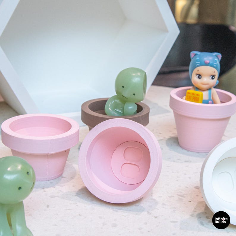 May include: A collection of small, pastel-colored flower pots in pink, brown, and white. Two green figurines and one doll are inside the pots. The doll is wearing a blue hat and overalls. The pots are arranged on a light-colored surface.