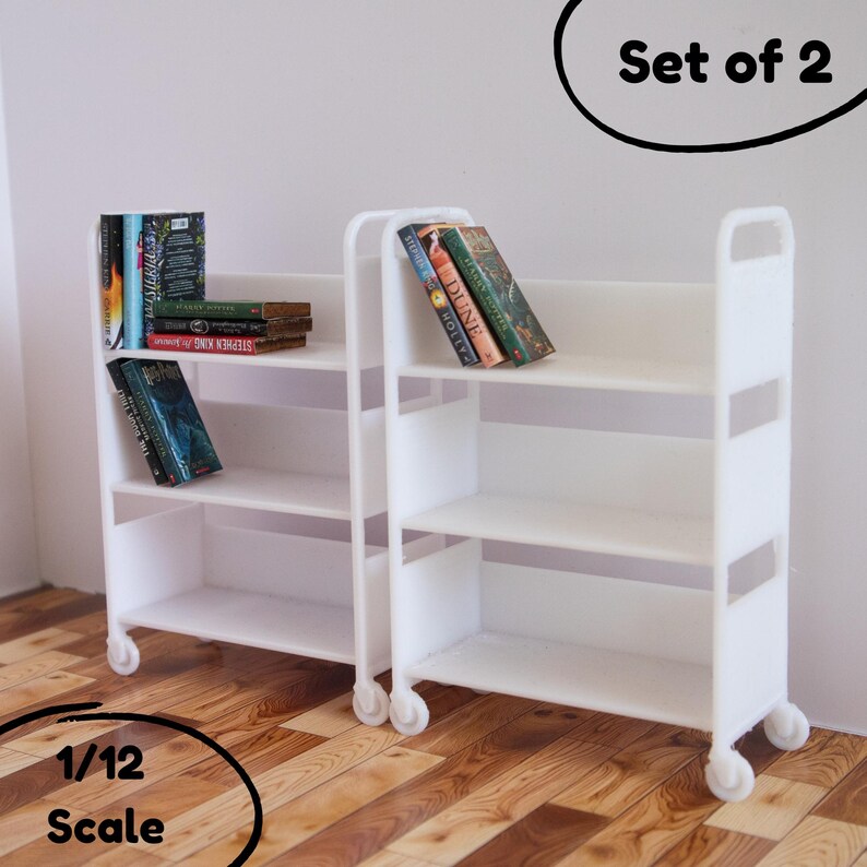 May include: Set of two white miniature bookcases with three shelves each. The bookcases are on wheels and have books on the shelves. The text "Set of 2" is written in black on a white circle above the bookcases. The text "1/12 Scale" is written in black on a white circle below the bookcases.