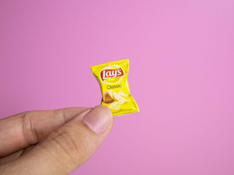 May include: A miniature yellow Lay's Classic potato chip bag with the brand's logo and the word "Classic" printed on it. The bag contains images of potato chips. The bag is held against a pink background.