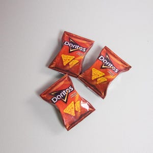 May include: Three bags of Doritos Nacho Cheese tortilla chips are arranged on a light gray surface. The bags are red and orange with the Doritos logo and images of the chips. The bags are slightly angled.
