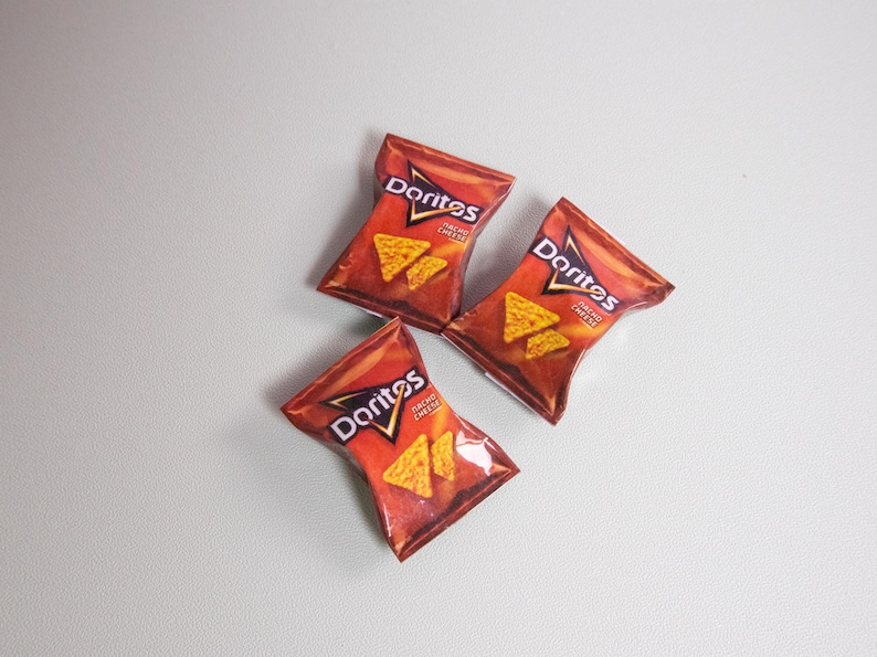 May include: Three bags of Doritos Nacho Cheese tortilla chips are arranged on a light gray surface. The bags are red and orange with the Doritos logo and images of the chips. The bags are slightly angled.