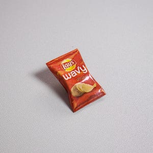 May include: A miniature bag of Lay's Wavy potato chips in a red and yellow package. The bag features the Lay's logo and the words "Wavy Original". Two wavy potato chips are visible on the front of the bag.