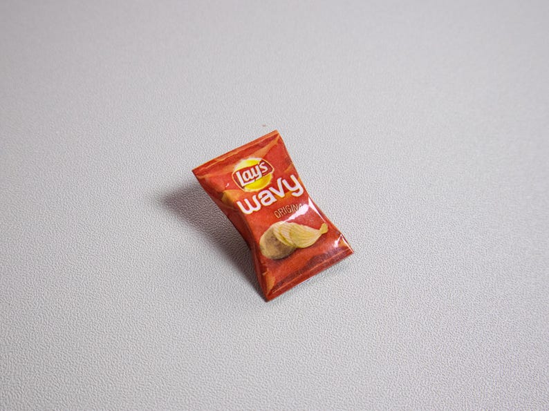 May include: A miniature bag of Lay's Wavy potato chips in a red and yellow package. The bag features the Lay's logo and the words "Wavy Original". Two wavy potato chips are visible on the front of the bag.