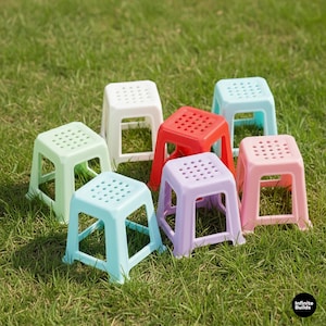 May include: A collection of seven colorful plastic stools in various shades, including red, pink, green, blue, white, and purple. Each stool has a square seat with small circular holes and a sturdy, open-frame base. The stools are arranged on a bed of green grass.