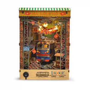K&ouml;nnte beinhalten: Ein detailliertes Buch-Eck aus Holz mit einer Zugszene in Hanoi, Vietnam. Das Diorama umfasst einen Zug, Backsteingeb&auml;ude und Lichterketten. Der Text "HA NOI PHO" und "PHO DUONG TAU" sind sichtbar.