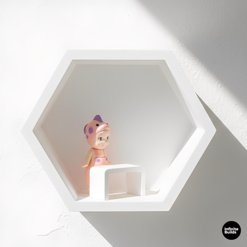 May include: A white, hexagonal wall shelf with a small, white, curved-leg table. A doll wearing a pink and purple spotted outfit and hat stands on the table. The doll has a serene expression. The shelf is mounted on a white wall.