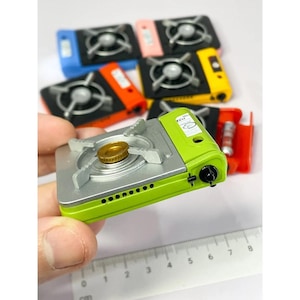 May include: A collection of miniature camping stoves in various colours, including green, yellow, blue, and red. The green stove is held in a hand, with a ruler in the foreground. The stoves have a silver burner and a gold nozzle.