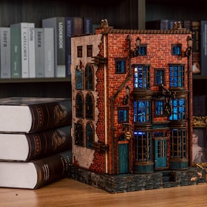 Pode incluir: Um modelo detalhado de um edif&iacute;cio em miniatura com fachada de tijolo, janelas iluminadas em azul e apar&ecirc;ncia desgastada. O modelo est&aacute; sobre uma superf&iacute;cie de madeira, perto de uma pilha de livros e uma foto emoldurada.