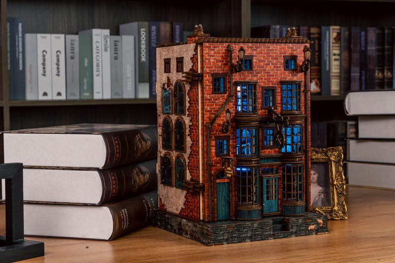 Pode incluir: Um modelo detalhado de um edif&iacute;cio em miniatura com fachada de tijolo, janelas iluminadas em azul e apar&ecirc;ncia desgastada. O modelo est&aacute; sobre uma superf&iacute;cie de madeira, perto de uma pilha de livros e uma foto emoldurada.