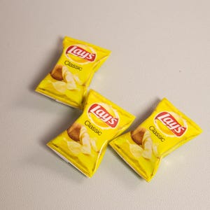 May include: Three yellow bags of Lay's Classic potato chips are arranged on a light gray surface. Each bag features the Lay's logo in red and white, with the word "Classic" below. The bags display images of potato chips.