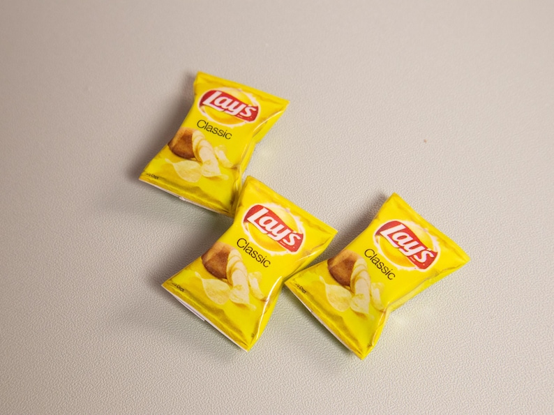 May include: Three yellow bags of Lay's Classic potato chips are arranged on a light gray surface. Each bag features the Lay's logo in red and white, with the word "Classic" below. The bags display images of potato chips.