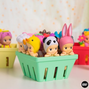 Puede incluir: Una colecci&oacute;n de peque&ntilde;as y coloridas figuras en una cesta verde. Las figuras est&aacute;n vestidas con varios disfraces de animales y alimentos, incluyendo un panda, un pato y un conejo. Otras cestas con figuras similares est&aacute;n en el fondo.