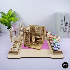 Può includere: Una stazione organizzatrice per il fai-da-te in legno chiaro con un diorama di una stanza in miniatura completo di cavalletto, scrivania e mobili. La stazione include fori circolari per barattoli di vernice e attrezzi, un bicchiere di plastica.
