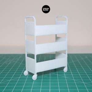 May include: White metal rolling cart with three shelves. The cart has four wheels and is on a green grid surface.