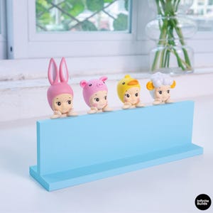 May include: A light blue display shelf with four small figurines. The figurines are dressed as a bunny, pig, duck, and sheep. The bunny is pink, the pig is pink, the duck is yellow, and the sheep is white with yellow accents.