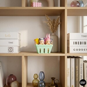 Puede incluir: Una cesta de pl&aacute;stico verde claro llena de peque&ntilde;as figuras de animales y personas. Las figuras incluyen un panda, un pato y una persona con un sombrero de conejo rosa. La cesta est&aacute; sobre una estanter&iacute;a de madera con libros y objetos decorativos.