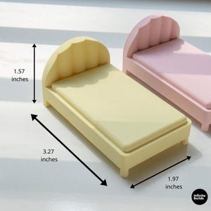 K&ouml;nnte beinhalten: Ein Miniaturbett in einem sanften Gelbton mit einem wellenf&ouml;rmigen Kopfteil. Das Bett misst 8,3 cm lang, 5 cm breit und 4 cm hoch. Ein weiteres Bett in Rosa ist im Hintergrund zu sehen. Die Betten sind f&uuml;r ein Puppenhaus oder eine Miniaturumgebung konzipiert.