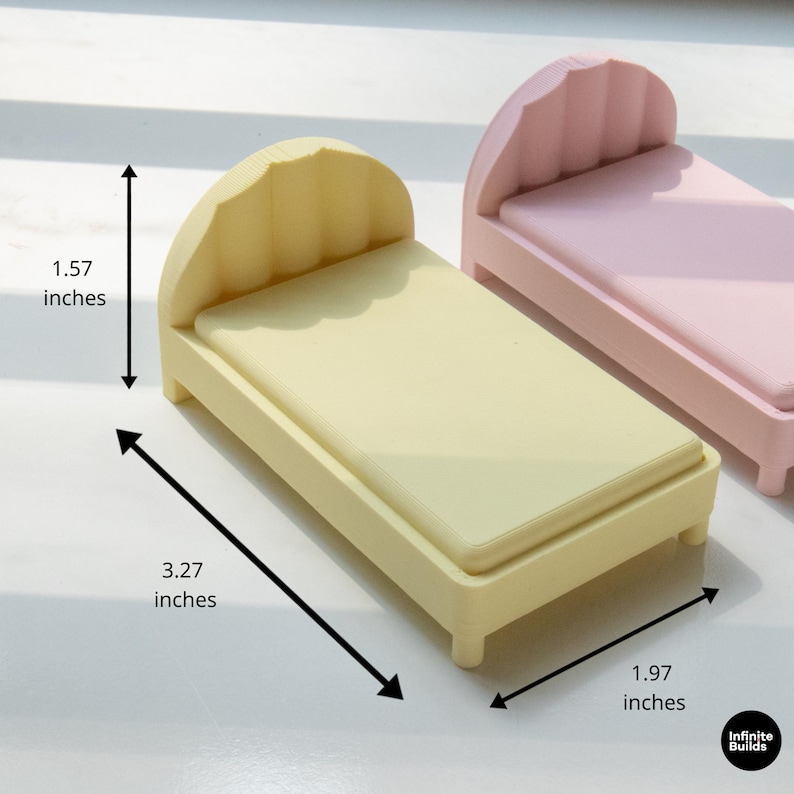 K&ouml;nnte beinhalten: Ein Miniaturbett in einem sanften Gelbton mit einem wellenf&ouml;rmigen Kopfteil. Das Bett misst 8,3 cm lang, 5 cm breit und 4 cm hoch. Ein weiteres Bett in Rosa ist im Hintergrund zu sehen. Die Betten sind f&uuml;r ein Puppenhaus oder eine Miniaturumgebung konzipiert.