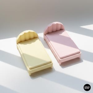 K&ouml;nnte beinhalten: Zwei Miniaturbetten, eines in blassem Gelb und eines in Rosa, werden gezeigt. Jedes Bett hat ein wellenf&ouml;rmiges Kopfteil und eine rechteckige Matratze. Die Betten sind klein, wahrscheinlich f&uuml;r Puppen oder Dekorationszwecke. Die Betten sind aus einem festen Material.