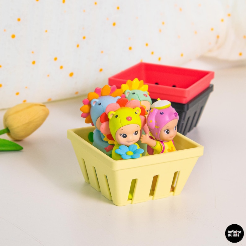Puede incluir: Una colecci&oacute;n de figuras coloridas en una cesta amarilla, con una cesta roja y negra en el fondo. Las figuras tienen varios sombreros con temas de animales y detalles de flores. El fondo incluye una superficie blanca y una almohada blanca con lunares naranjas.