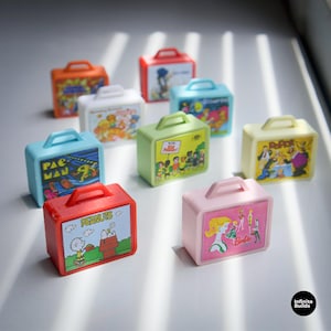May include: A collection of miniature, colorful lunchboxes. Each box features a different cartoon or character design, including Pac-Man, Barbie, and Peanuts. The boxes have a rectangular shape with a handle on top.