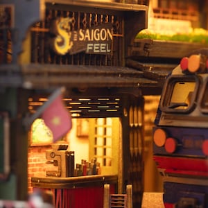 K&ouml;nnte beinhalten: Detailliertes Miniaturmodell einer vietnamesischen Stra&szlig;enszene mit einem Gesch&auml;ft mit dem Schild "SAIGON FEEL". Das Modell umfasst eine kleine Flagge, eine Bar und einen Zugwagen. Die Szene ist mit warmem, einladendem Licht beleuchtet.
