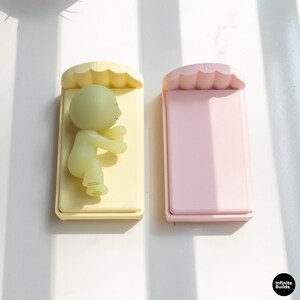 K&ouml;nnte beinhalten: Zwei rechteckige Notizbl&ouml;cke in Pastellfarben. Einer ist gelb mit einer kleinen, gr&uuml;nen Figur einer Person, die darauf liegt. Der andere ist rosa. Beide haben einen wellenf&ouml;rmigen oberen Rand. Die Bl&ouml;cke befinden sich auf einer wei&szlig;en Oberfl&auml;che mit Schatten.