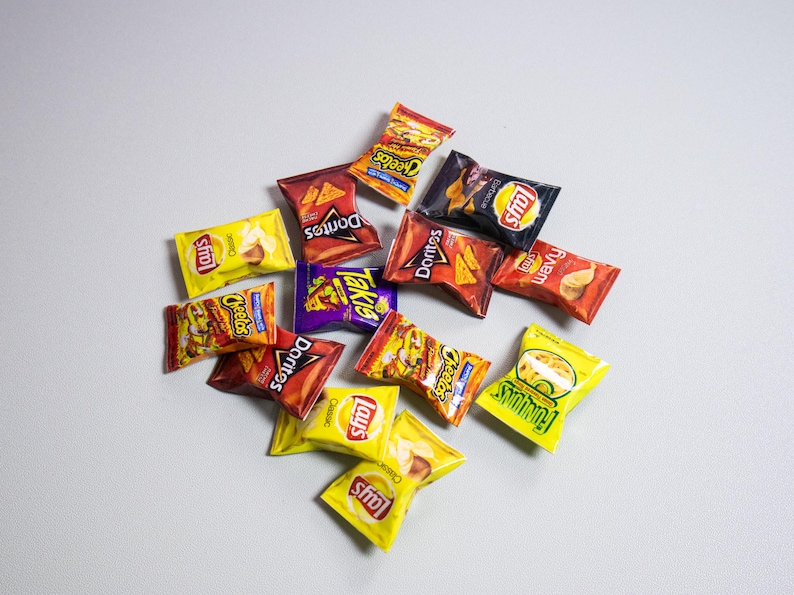 May include: A collection of various snack-sized chip bags, including Doritos, Cheetos, Lays, and Takis, in a variety of colors. The bags are scattered on a light gray surface, showcasing the different brands and flavors.