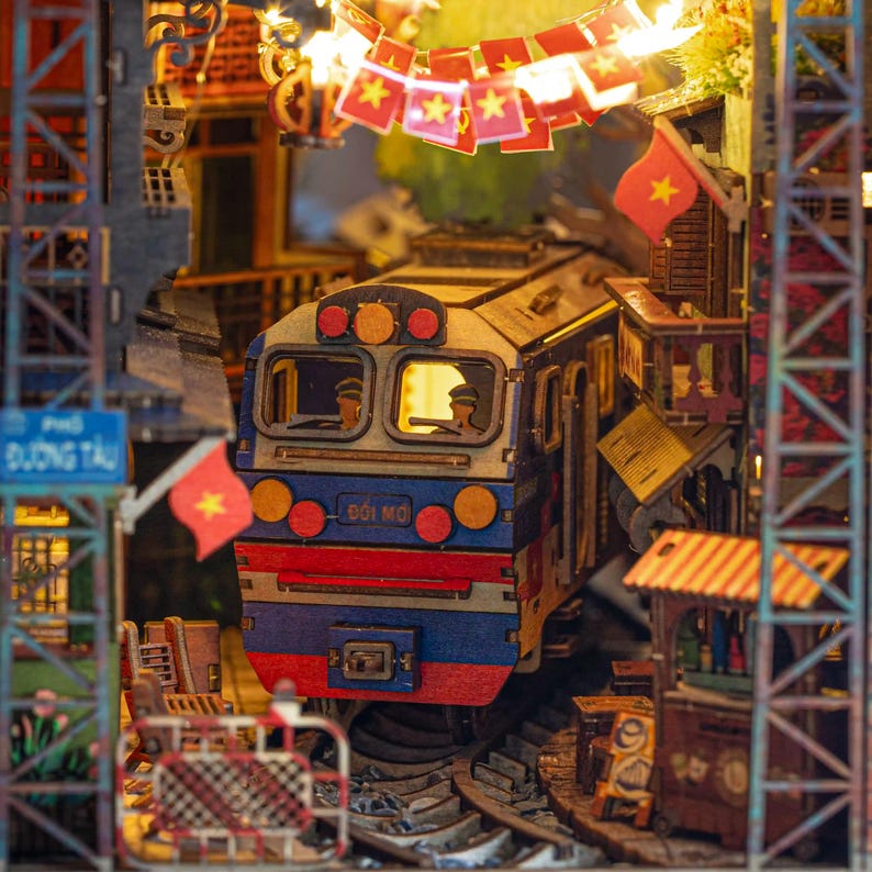 K&ouml;nnte beinhalten: Detailliertes Holzmodell einer Bahnhofsszene. Der Zug ist blau, rot und wei&szlig;, mit dem Text "DOI MO" auf der Vorderseite. Die Szene umfasst Miniaturflaggen, Geb&auml;ude und ein Schild mit der Aufschrift "PHO DUONG TAU".
