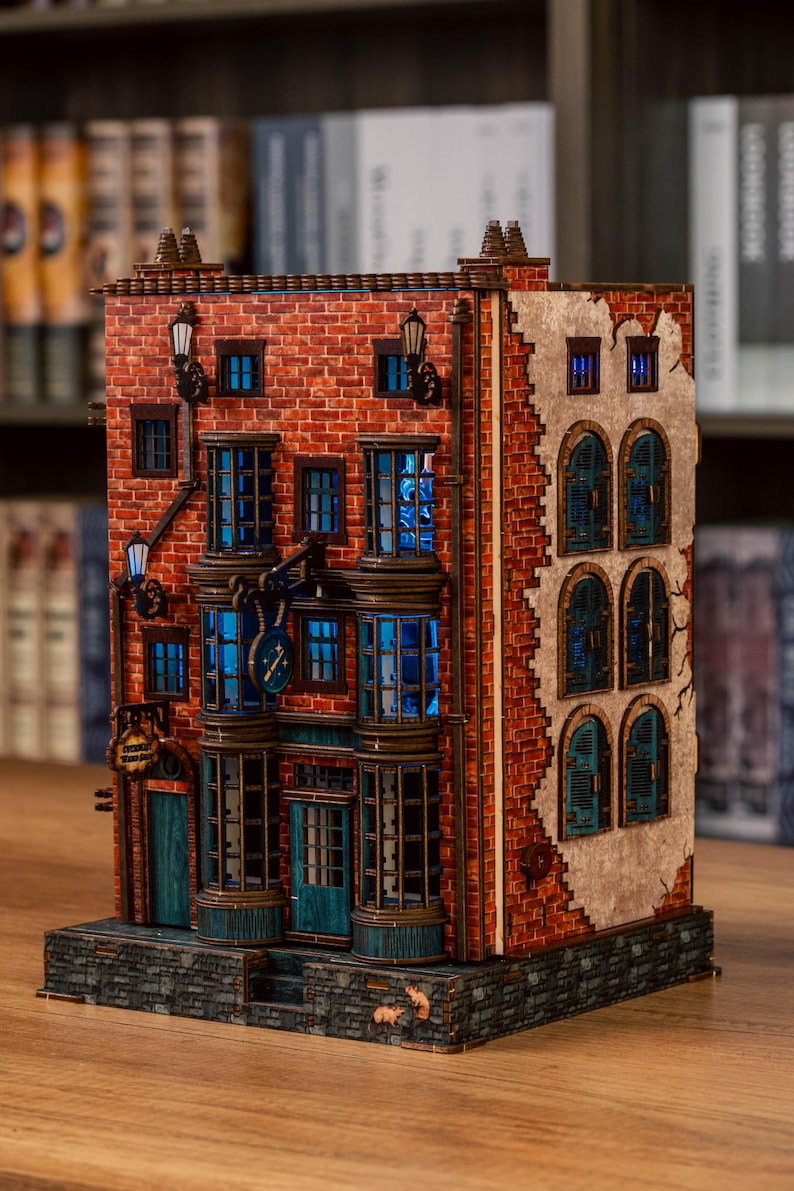 Pode incluir: Modelo de madeira detalhado de um edif&iacute;cio com fachada de tijolo, janelas azuladas e l&acirc;mpadas decorativas. O modelo apresenta um rel&oacute;gio e um letreiro acima da entrada. O interior do edif&iacute;cio &eacute; iluminado por luz azul.