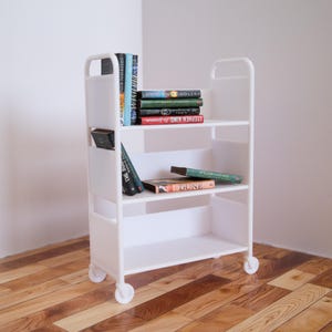 May include: White rolling bookshelf with four shelves, filled with books. The top shelf has a stack of books with the title "STEPHEN KING" visible. The second shelf has a stack of books with the title "THE DUNE" visible. The bottom shelf is empty.