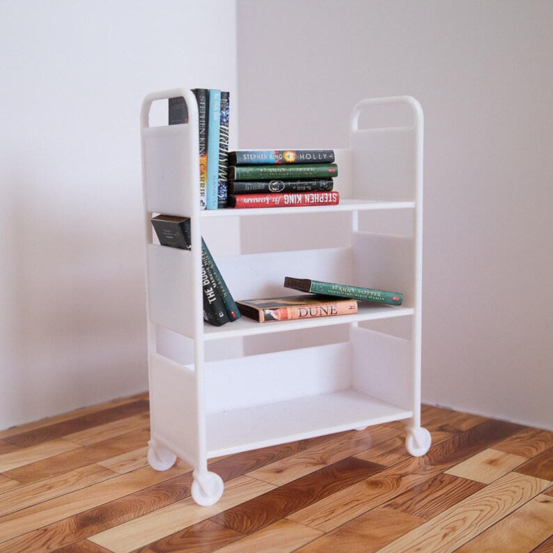 May include: White rolling bookshelf with four shelves, filled with books. The top shelf has a stack of books with the title "STEPHEN KING" visible. The second shelf has a stack of books with the title "THE DUNE" visible. The bottom shelf is empty.