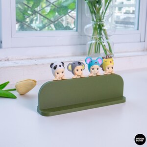 May include: A green shelf displays four small figurines, each wearing a different animal hat: panda, koala, mouse, and lion. The figurines have light skin and are positioned in a row. The shelf is on a white surface.