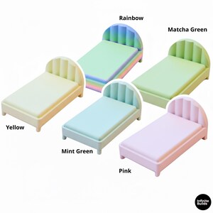 K&ouml;nnte beinhalten: Eine Sammlung von Miniatur-Betten in Pastellfarben: Gelb, Mintgr&uuml;n, Rosa, Matcha-Gr&uuml;n und Regenbogen. Jedes Bett hat ein abgerundetes Kopfteil mit vertikalen Rillen und eine rechteckige Matratze.