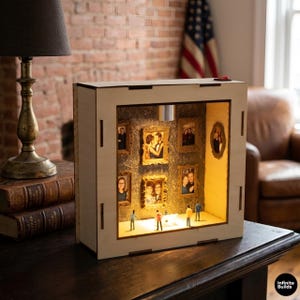 May include: A square light-colored wooden display box featuring an internal illuminated gallery. The interior walls are lined with patterned wallpaper and decorated with several small gold-toned picture frames containing photographs.
