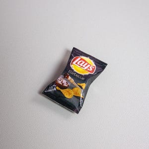 May include: A miniature bag of Lay's Barbecue potato chips. The bag is dark with the Lay's logo in red and white on a yellow circle. The word "Barbecue" is printed below the logo, with an image of chips and sauce.