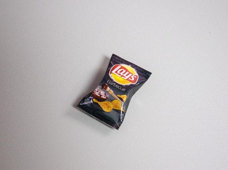May include: A miniature bag of Lay's Barbecue potato chips. The bag is dark with the Lay's logo in red and white on a yellow circle. The word "Barbecue" is printed below the logo, with an image of chips and sauce.