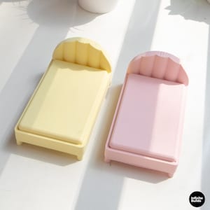 K&ouml;nnte beinhalten: Zwei Miniatur-Organizer in Bettform, einer hellgelb und einer rosa. Jeder hat ein gewelltes Kopfteil und eine rechteckige Basis. Die Betten sind f&uuml;r kleine Gegenst&auml;nde konzipiert, mit einer minimalistischen &Auml;sthetik.