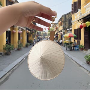 May include: A woven, beige conical hat keychain with a gold-colored clasp and a brown leather strap. The hat is held up in front of a street scene with yellow buildings and colorful lanterns.